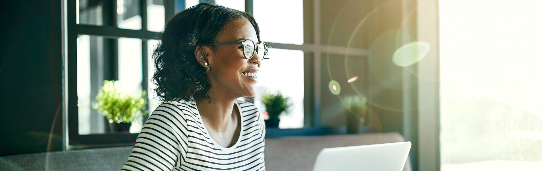 A smiling woman in front of a laptop in a sunny room.