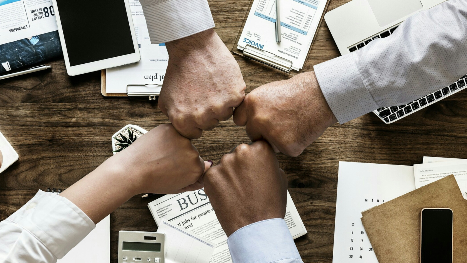 Four hands doing knuckle bumps above table with devices and business documents.